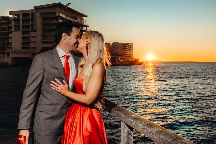 The couple smiles at each other with the sunset and ocean behind them, creating a warm glow.