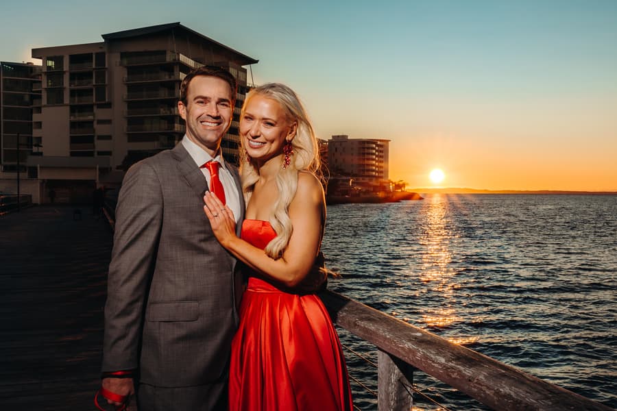 The couple stands on Woody Point Jetty at sunset, smiling warmly. The sun sets over the water, casting a golden glow.