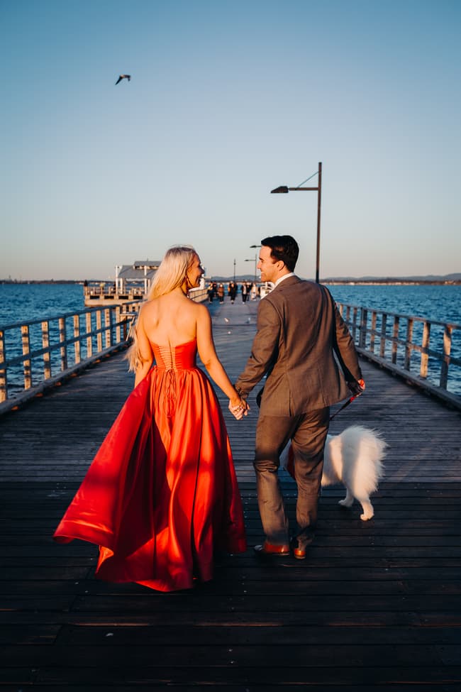 The couple walks hand in hand along the jetty, accompanied by a fluffy white dog. The sky is clear and blue.