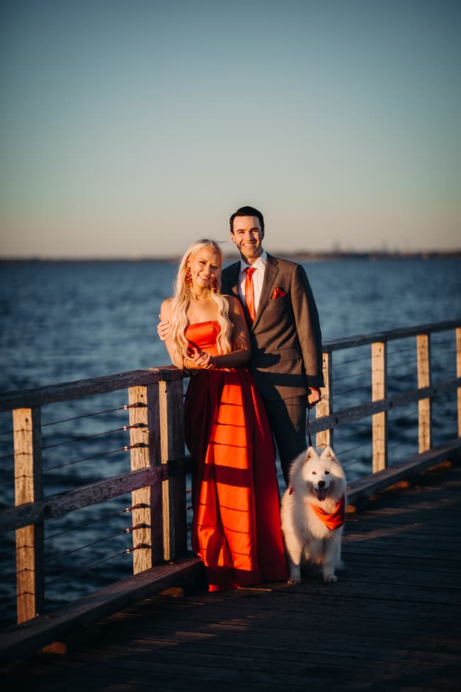 The couple poses with their dog on the jetty, the ocean in the background. They are smiling and relaxed.