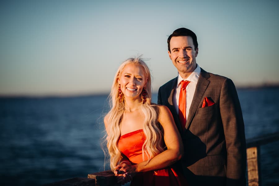The couple stands close together on the jetty, smiling at the camera with the ocean behind them.
