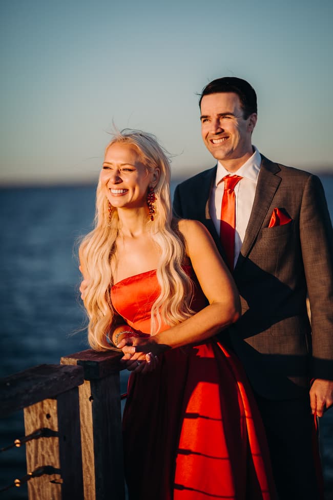 The couple leans on the railing of the jetty, gazing out at the ocean with happy expressions.
