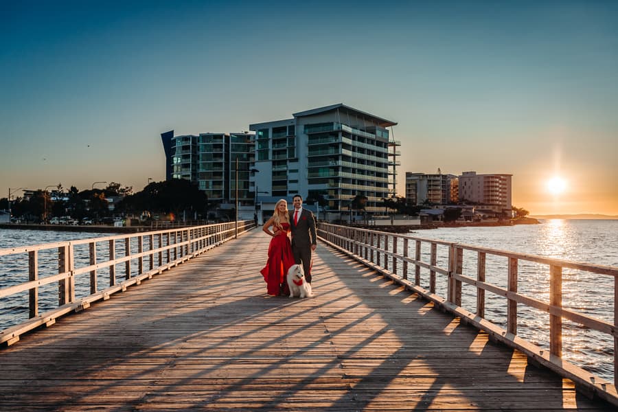 The couple stands on the jetty with their dog, the sun setting behind them. They look content and happy.