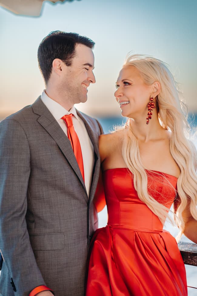 The couple stands on the jetty, facing each other with smiles. The ocean and sky are a brilliant blue.