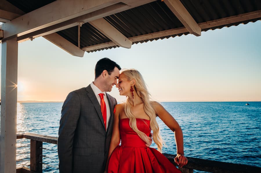 The couple stands under a shelter on the jetty, leaning in close with the ocean in the background.