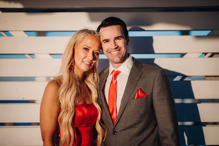 The couple stands together, smiling warmly at the camera. The ocean is visible behind them.