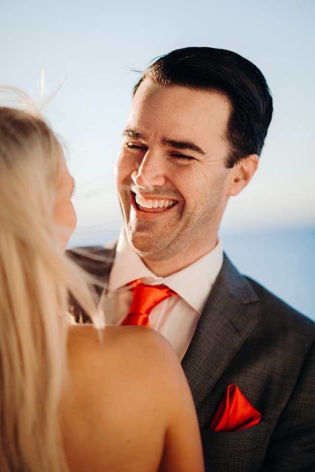 The couple shares a light moment, laughing together. The blue sky and ocean are in the background.