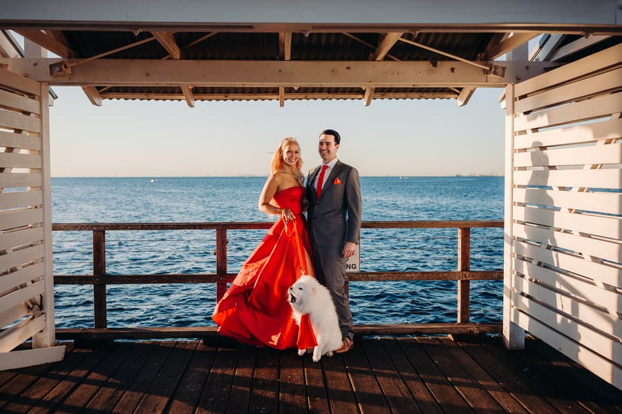 The couple stands on the jetty with their dog, framed by the ocean and sky. They look happy and relaxed.