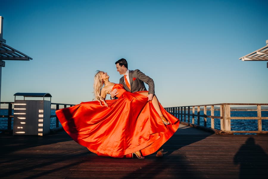 The couple shares a playful dance on the jetty, the woman's dress flowing dramatically. The ocean is behind them.