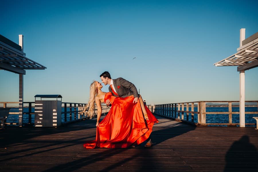 The couple dances on Woody Point Jetty, with the person in a red dress being dipped by the other in a suit. The sun casts long shadows.