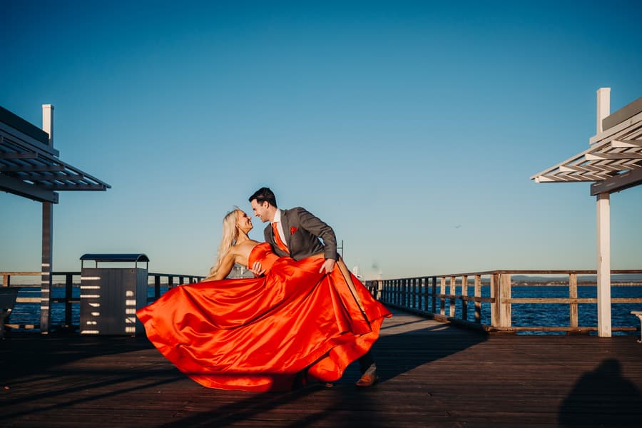 The couple twirls on Woody Point Jetty, the red dress flowing elegantly as they move. The sky is clear and blue.