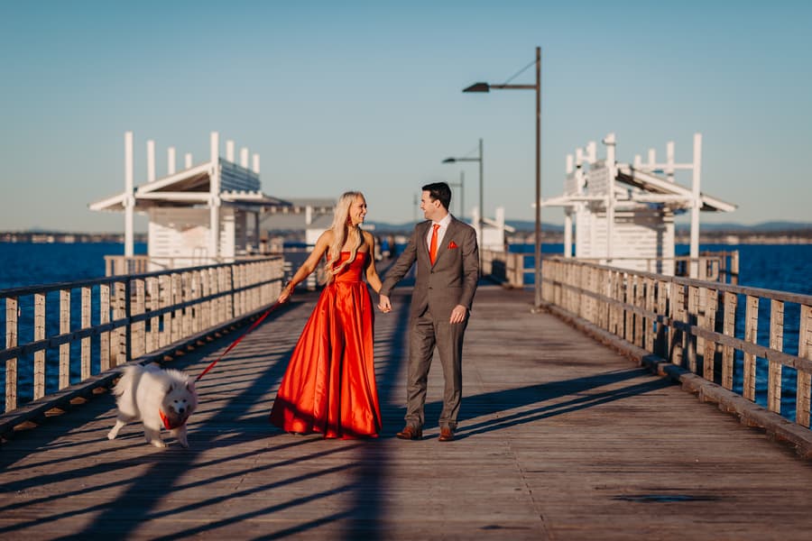 The couple walks hand in hand on Woody Point Jetty, with a fluffy white dog on a leash beside them.