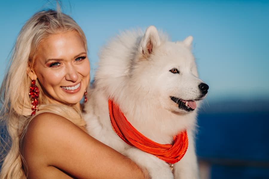 A person in a red dress holds a fluffy white dog with an orange bandana, both smiling at the camera.
