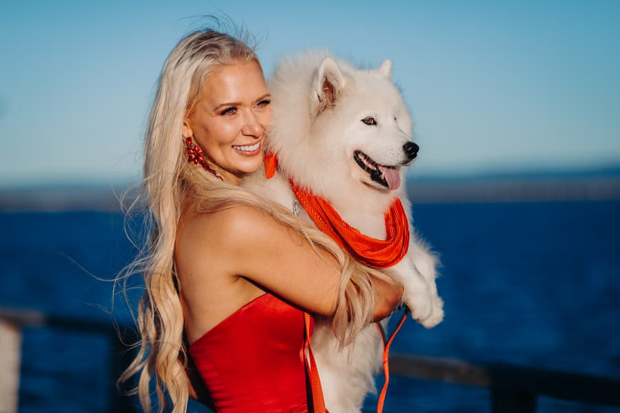 A person in a red dress holds a fluffy white dog against a backdrop of blue water, both looking content.