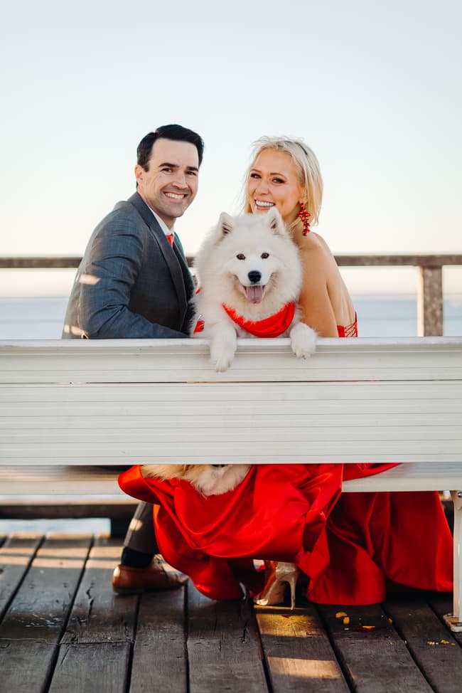 The couple sits on a bench with their fluffy white dog between them, all looking at the camera.