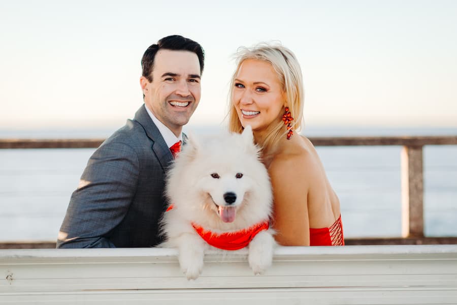 The couple and their fluffy white dog sit on a bench, smiling warmly at the camera.