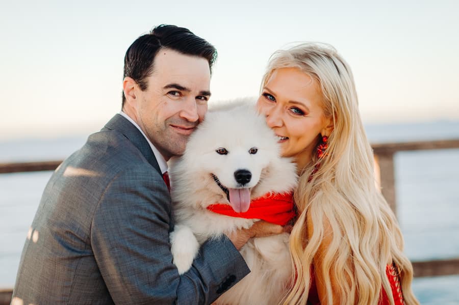 The couple embraces their fluffy white dog, all three looking happy and close on the jetty.