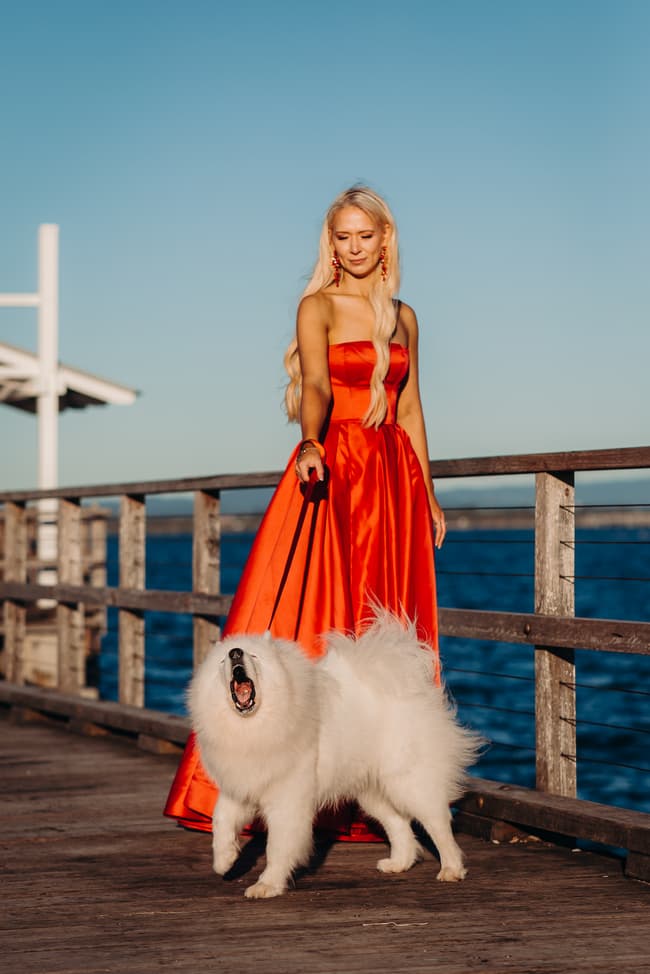 Person in a red dress walking a fluffy white dog on Woody Point Jetty, with the ocean in the background.