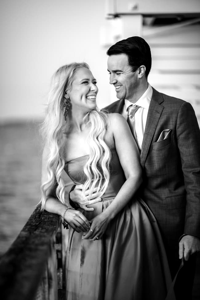 Black and white image of a couple laughing together on Woody Point Jetty, with the ocean in the background.