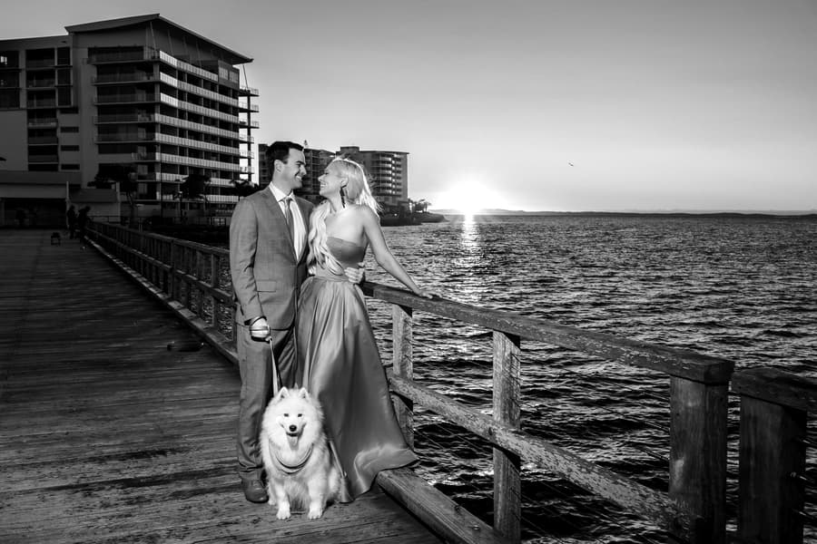 Black and white photo of a couple and a dog on Woody Point Jetty, with the sun setting over the water.