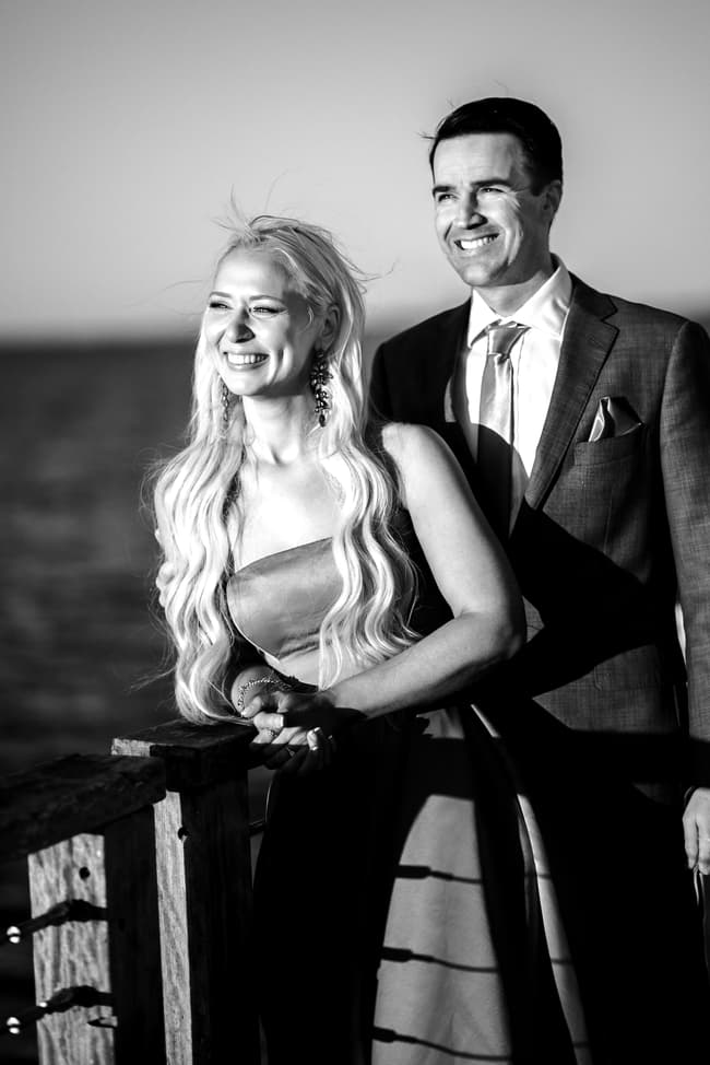 Black and white image of a couple smiling and leaning on a railing at Woody Point Jetty, with the ocean behind them.