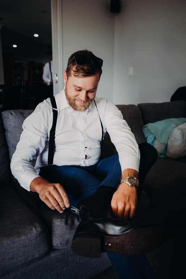 The groom Trent sits on a gray couch at Ocean View Estates, wearing a white shirt with black suspenders and blue pants, smiling as he adjusts his black boot.