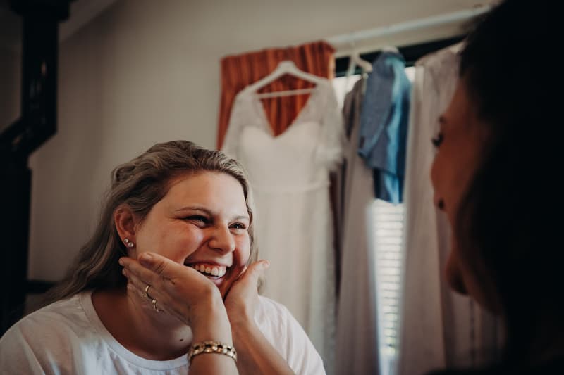 The bride Annie smiles as another woman gently holds her face, with a wedding dress hanging in the background at Ocean View Estates.