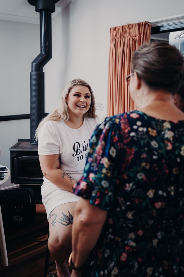 The bride Annie sits on a stool indoors wearing a white 'Bride to Be' t-shirt and shorts, smiling and talking to an older woman in a floral dress at Ocean View Estates.