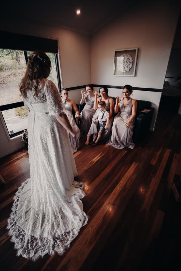 The bride Annie in a lace wedding gown stands facing four bridesmaids in matching dresses seated on a black couch at Ocean View Estates. A young boy in suspenders sits on one bridesmaid's lap. The group is indoors near a window with wooden floors.