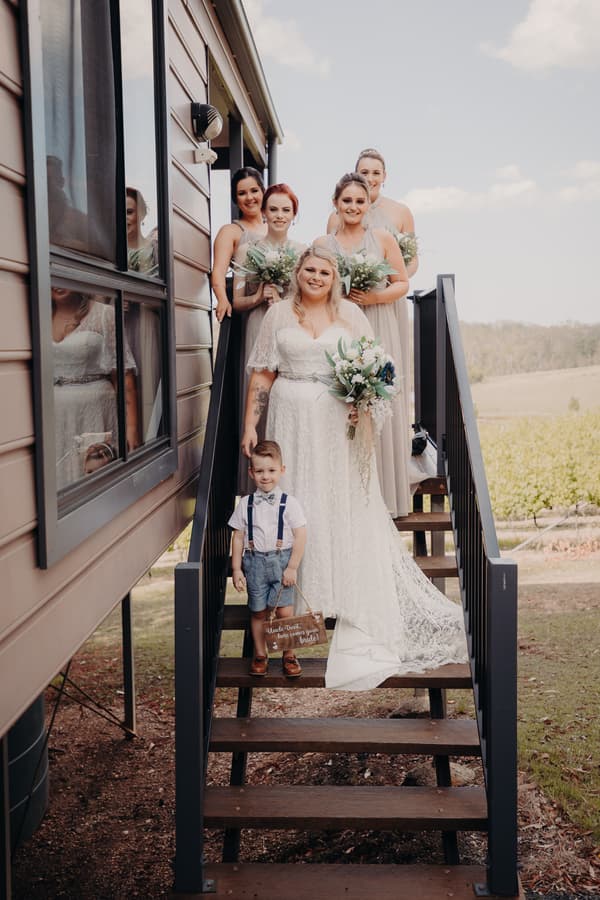 The bride Annie stands on outdoor stairs at Ocean View Estates holding a bouquet, accompanied by a young boy holding a sign and four bridesmaids in matching dresses holding bouquets behind her.