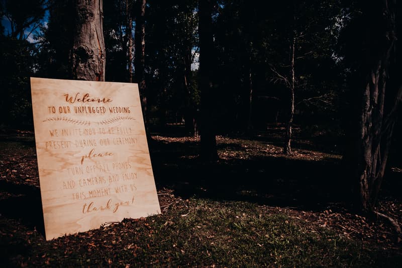 Wooden sign at Ocean View Estates reading 'Welcome to our unplugged wedding. We invite you to be fully present during our ceremony. Please turn off all phones and cameras and enjoy this moment with us. Thank you!' placed outdoors among trees and grass.