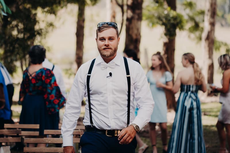 The groom Trent wearing a white shirt with black suspenders and a white bow tie walks outdoors at Ocean View Estates with guests mingling in the background.