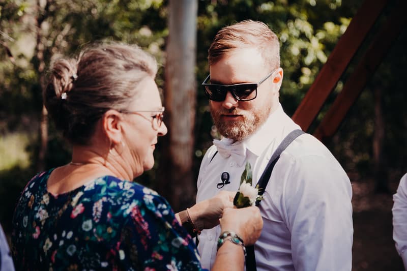 The groom Trent wearing sunglasses and a white shirt with suspenders has a boutonniere pinned on by an older woman, likely the mother of the groom, at Ocean View Estates.