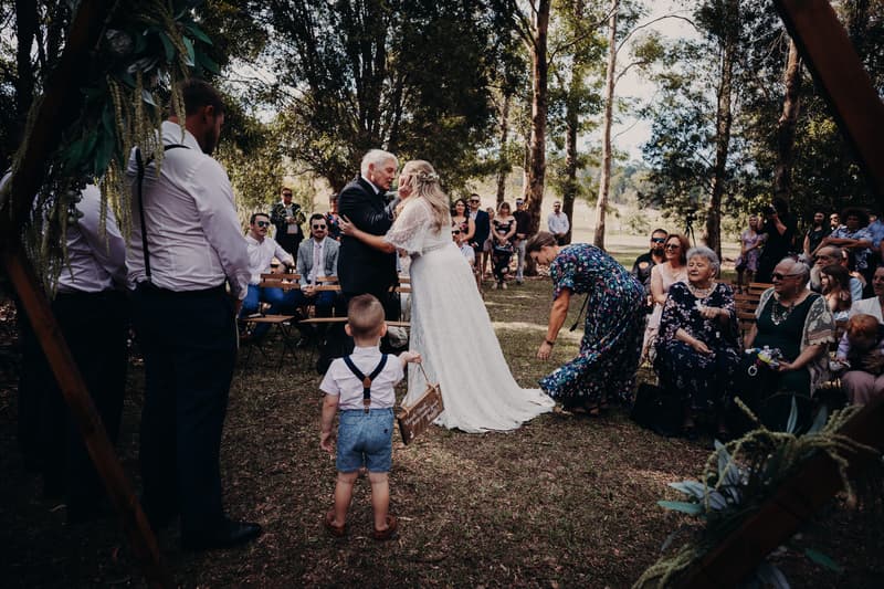 The bride Annie embraces an older man, likely her father, at Ocean View Estates during the ceremony while a young boy holding a wooden sign stands nearby and guests watch seated on benches outdoors.