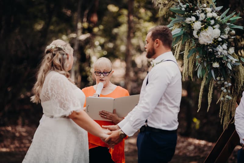Bride Annie and groom Trent hold hands facing each other during their wedding ceremony at Ocean View Estates, while an officiant reads from a book into a microphone.
