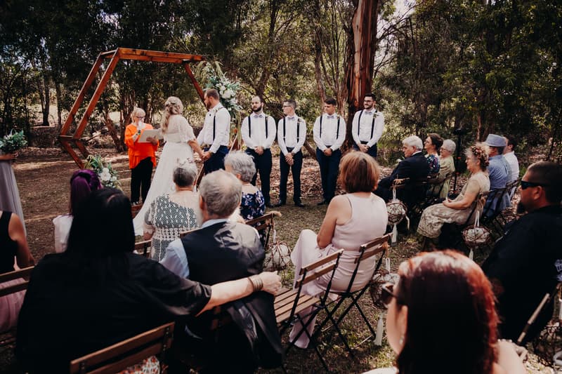 The bride Annie and groom Trent stand holding hands at the ceremony altar at Ocean View Estates, with the officiant reading from a book. Five groomsmen stand to the right of the couple, and guests are seated on wooden chairs watching the ceremony outdoors among trees.