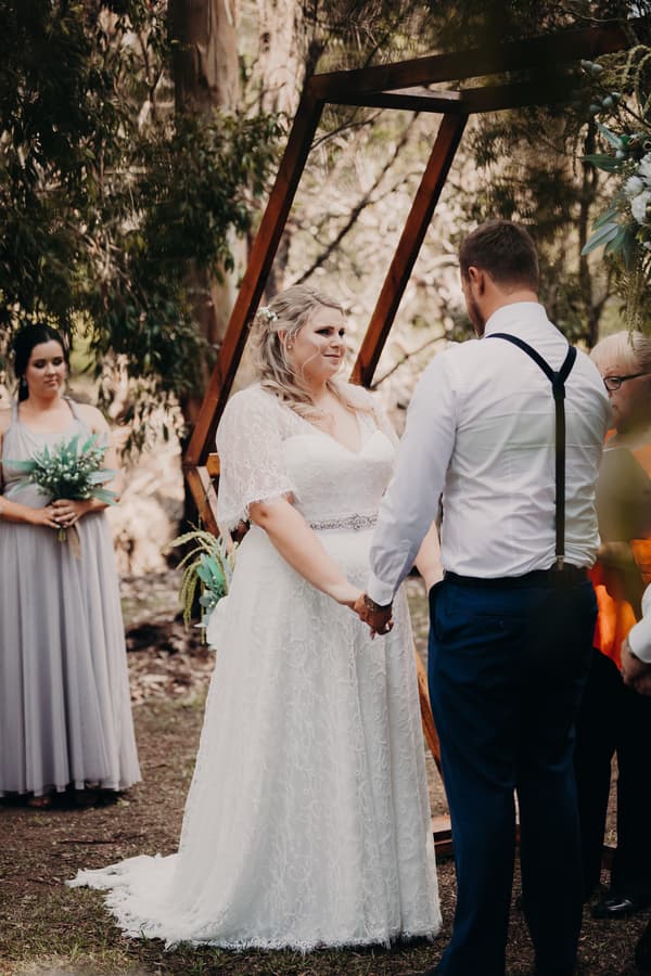 Bride Annie and groom Trent hold hands facing each other under a wooden arch at Ocean View Estates during their wedding ceremony, with a bridesmaid holding a bouquet standing to the side.