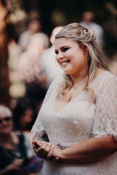 The bride Annie stands smiling in a white lace wedding dress, with blurred guests in the background. Brisbane videography by She Said Yes.