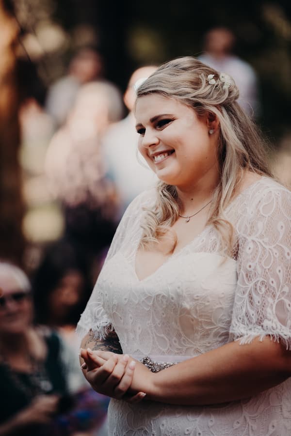 The bride Annie stands smiling in a white lace wedding dress at Ocean View Estates, with blurred guests in the background.
