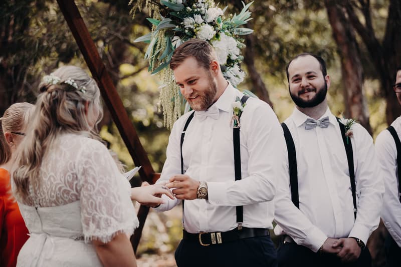 Annie and Trent exchange rings during their wedding ceremony at Ocean View Estates, with groomsmen standing nearby and floral decorations in the background.