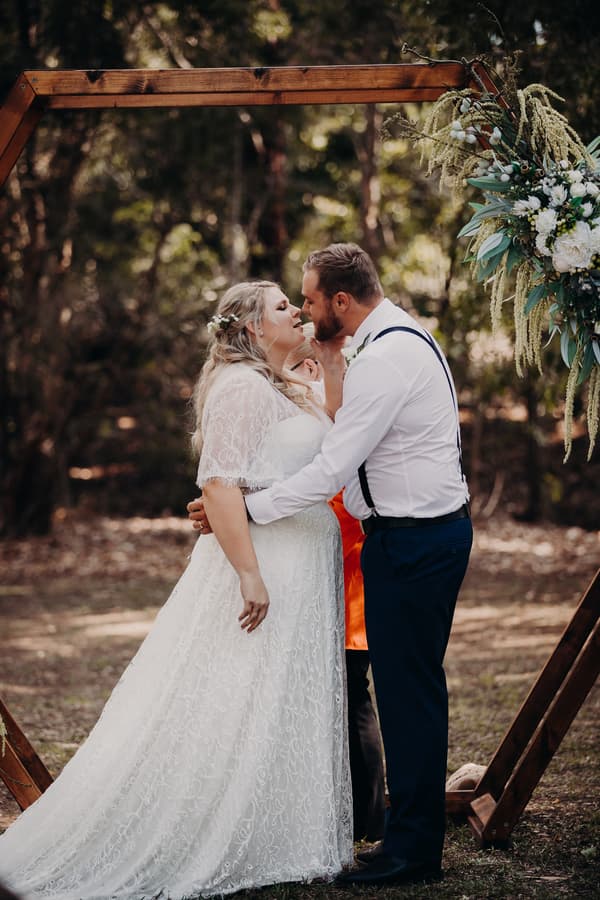 The bride Annie and groom Trent share a close moment at the ceremony stage under a wooden arch decorated with flowers at Ocean View Estates.