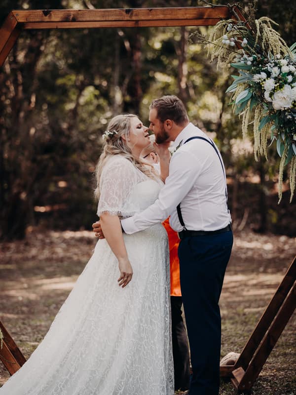 The bride Annie and groom Trent share a close moment at the ceremony stage under a wooden arch decorated with flowers at Ocean View Estates.