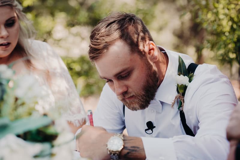 The groom Trent, wearing a white shirt with suspenders and a boutonniere, is signing a document at Ocean View Estates during the ceremony stage. The bride Annie is partially visible beside him.