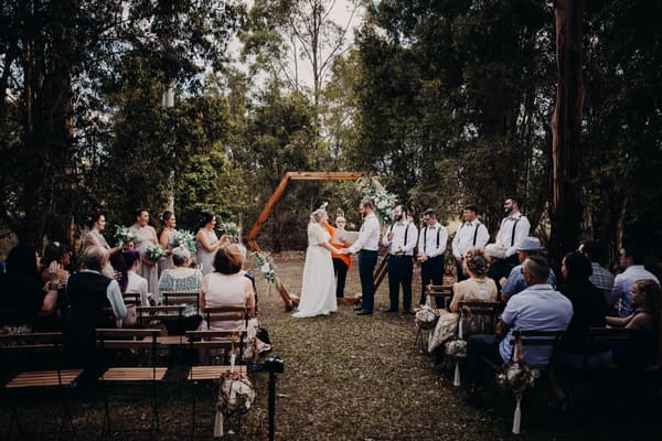 Bride Annie and groom Trent stand holding hands in front of an officiant under a wooden hexagonal arch decorated with flowers at Ocean View Estates, surrounded by bridesmaids, groomsmen, and seated guests during the wedding ceremony.