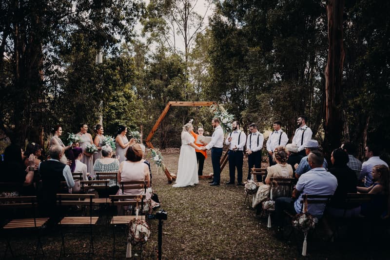 Bride Annie and groom Trent stand holding hands in front of an officiant under a wooden hexagonal arch decorated with flowers at Ocean View Estates, surrounded by bridesmaids, groomsmen, and seated guests during the wedding ceremony.