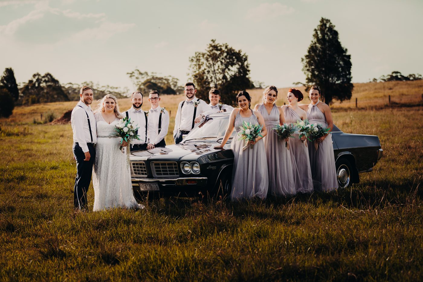 Annie the bride and Trent the groom stand with their bridal party beside a vintage black car in a grassy field. The bride holds a bouquet, and the bridesmaids wear matching lavender dresses holding bouquets. The groomsmen wear white shirts with suspenders and bow ties. Brisbane videography by She Said Yes.