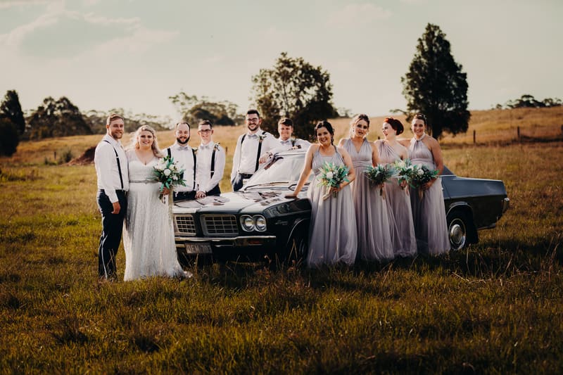 Annie the bride and Trent the groom stand with their bridal party beside a vintage black car in a grassy field. The bride holds a bouquet, and the bridesmaids wear matching lavender dresses holding bouquets. The groomsmen wear white shirts with suspenders and bow ties. Bridal editorial photography by She Said Yes.