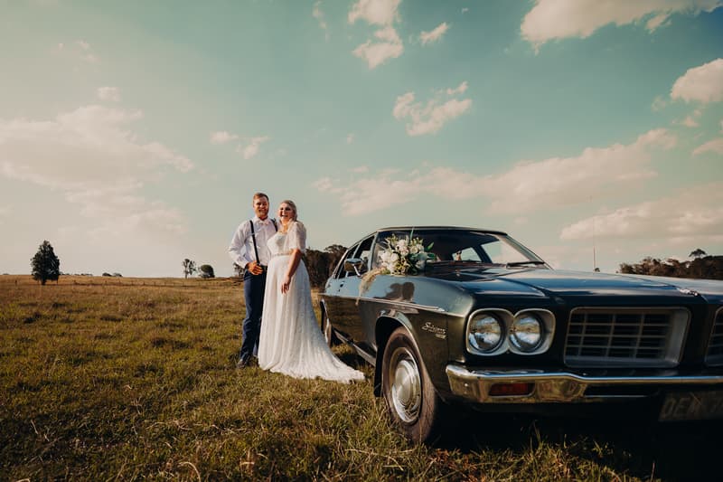 Annie the bride and Trent the groom stand together in a grassy field next to a vintage dark green car decorated with a floral arrangement at Ocean View Estates.