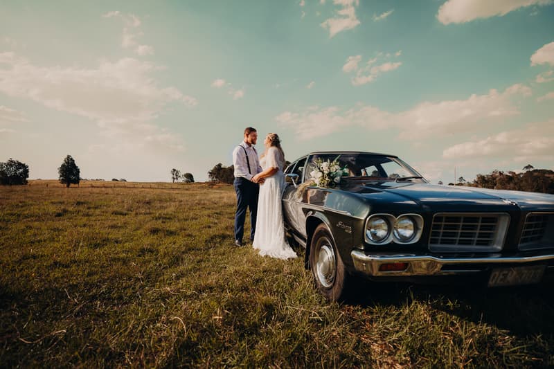 Annie the bride and Trent the groom stand holding hands beside a vintage dark green car with a floral bouquet on the hood at Ocean View Estates, in an open grassy field under a partly cloudy sky.