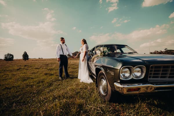 The bride Annie and groom Trent hold hands standing in a grassy field next to a vintage dark green car decorated with a white floral bouquet at Ocean View Estates.
